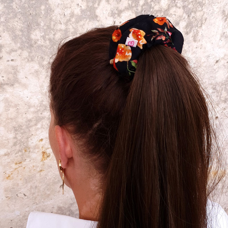 une femme a attaché ses cheveux en une queue de cheval avec un chouchou noir à fleurs orangées de taille médium de la marque Bunzelle